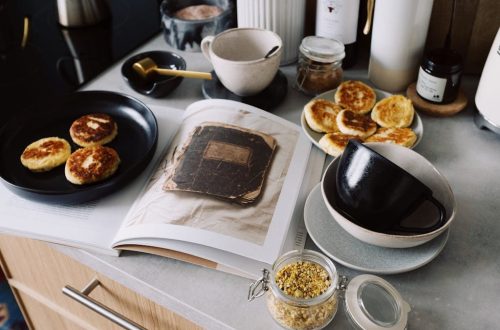 High angle of cookbook and cooked fritters on plates placed on table near utensils and ingredients in cozy kitchen at home